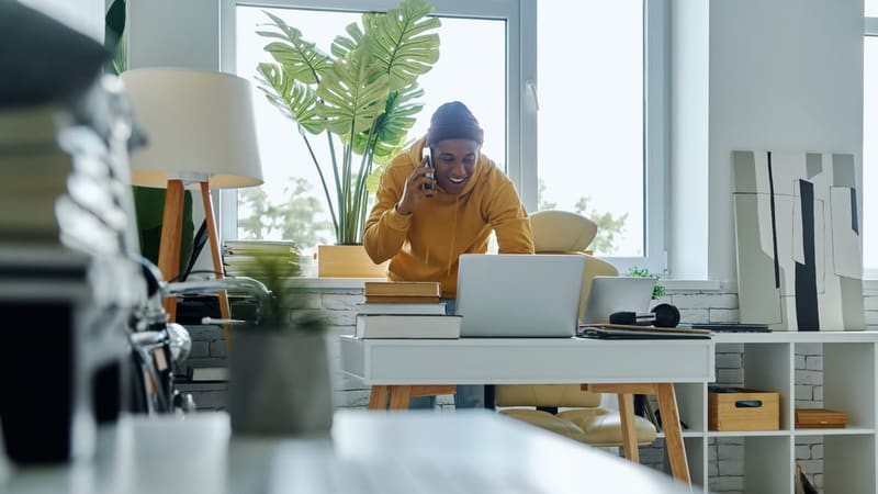 Un homme souriant en sweat jaune parle au téléphone devant son ordinateur portable dans un bureau lumineux